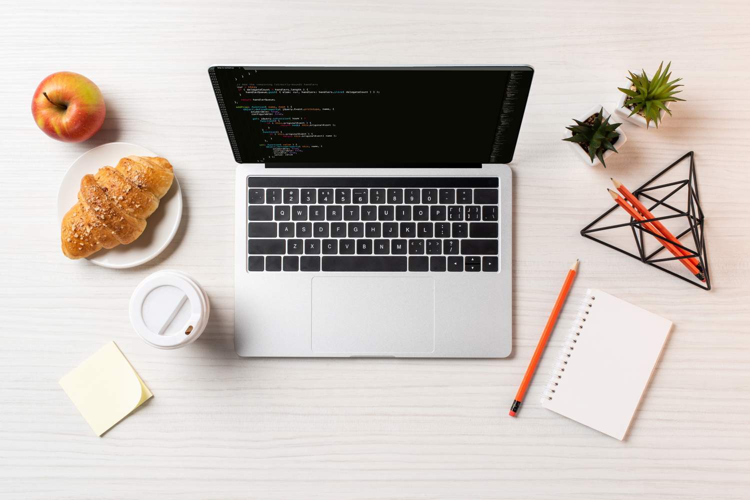 Work laptop, croissant, apple, two green plants, a coffee cup, a notepad and pens on a desk 