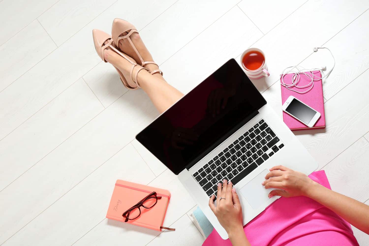 Woman sitting on the floor working on her Macbook with a notebook, tea cup and her phone beside her