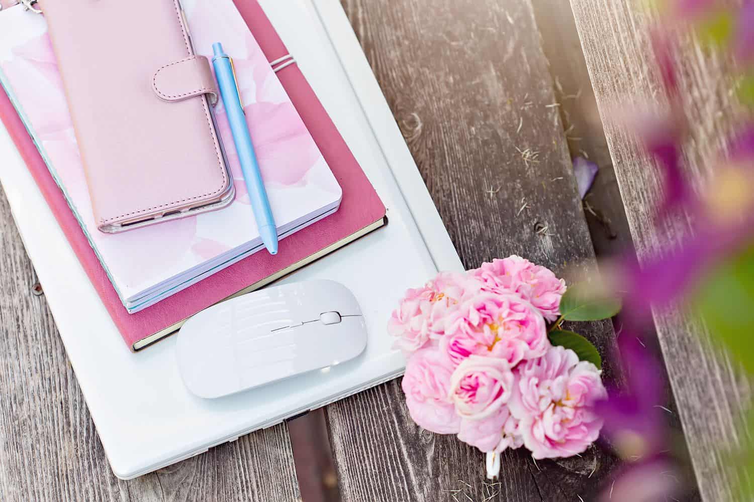 Pink flowers, computer mouse, notebooks, mobile phone and a blue pen on a brown bench