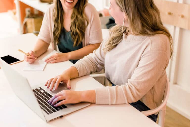 Two women sitting at a desk and talking, writing on a laptop