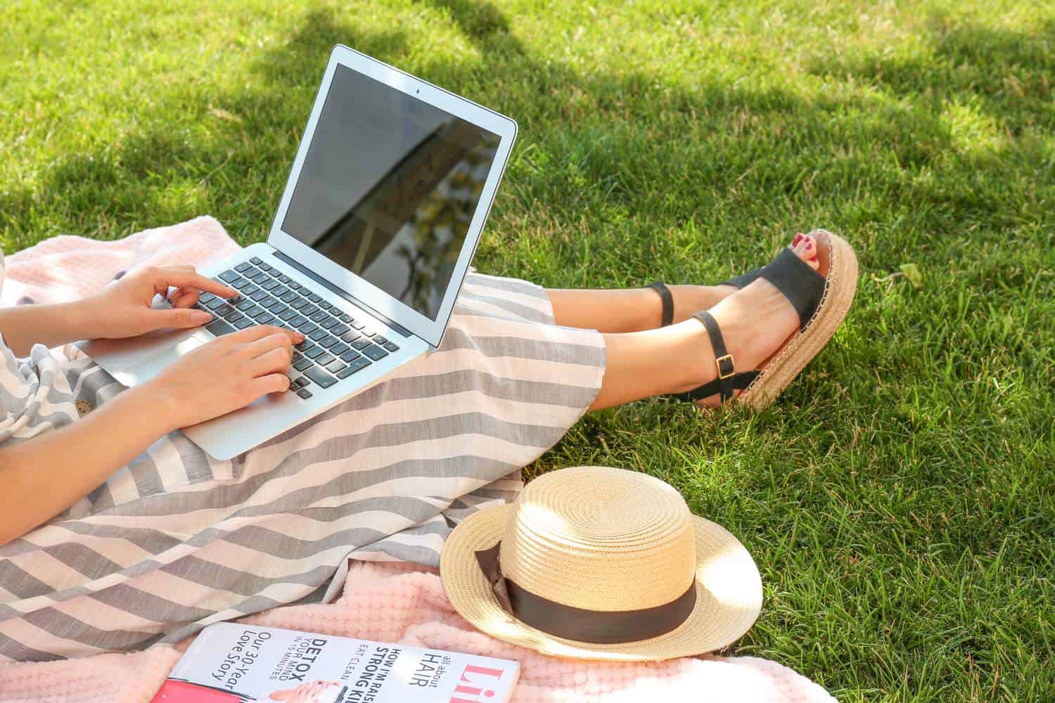 Woman working on a laptop in a green park with a summer hat
