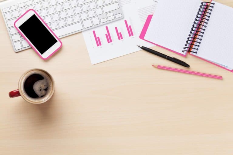 A pink phone, keyboard, coffee mug, notebook and a chart on an office desk