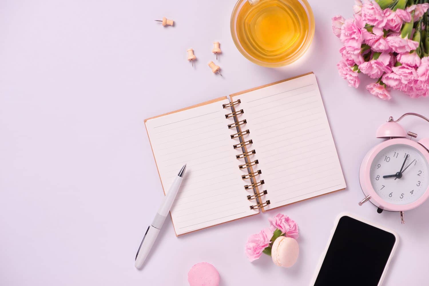 An open planner, pen, tea, an alarm clock and fresh flowers on a lilac background