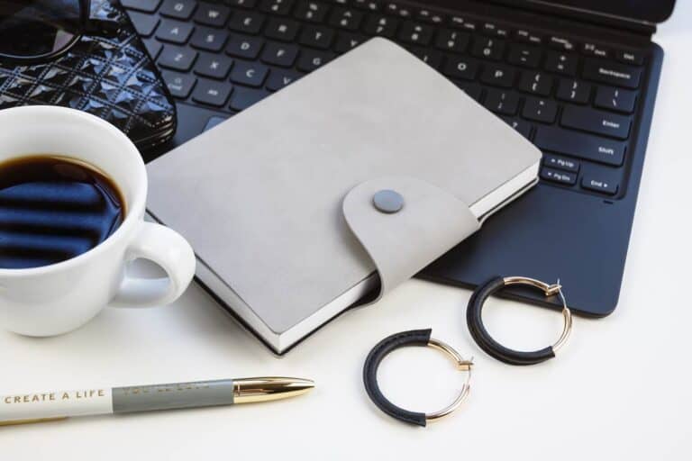 A gray notebook, black laptop, coffee cup, earrings and a pen that says "Create a life you'll love"