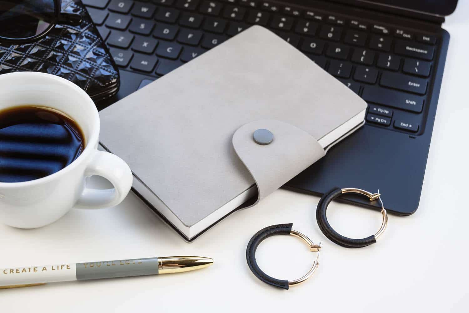 A gray notebook, black laptop, coffee cup, earrings and a pen that says "Create a life you'll love"
