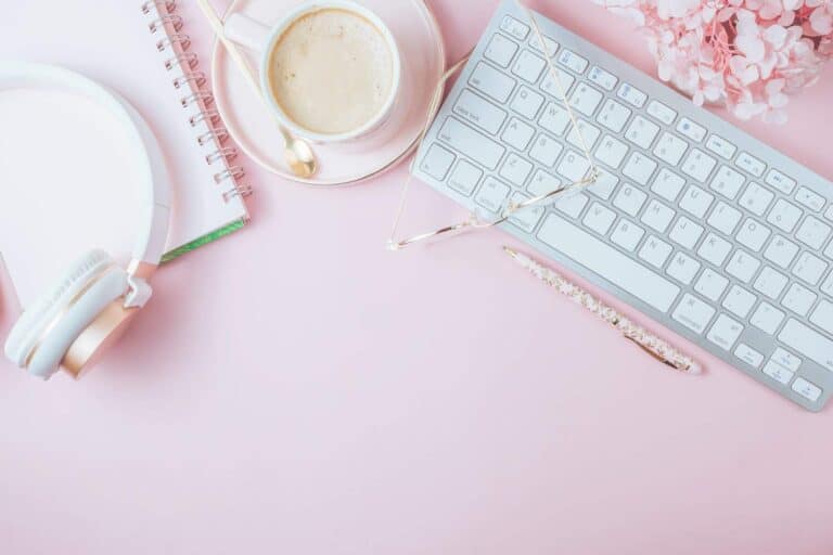 A leveling up setting with a white keyboard, coffee cup, notebook, golden phone and white headphones on a pink background