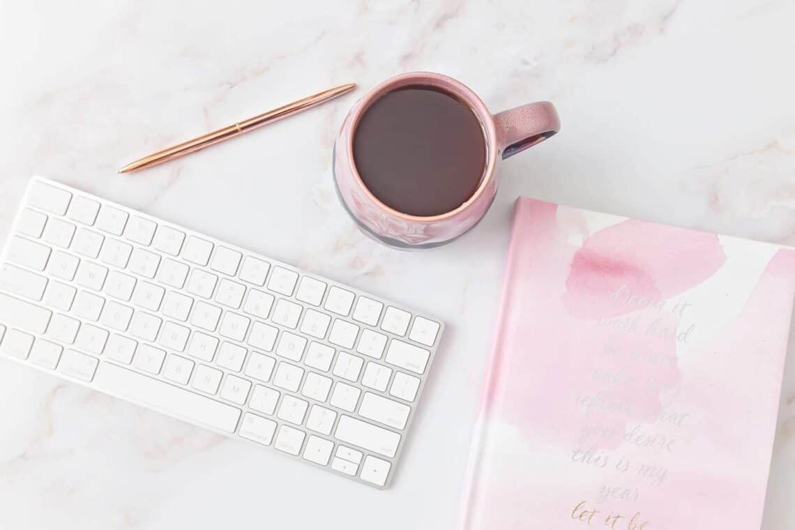 A beautiful pink and white notebook, coffee mug, keyboard an a pen on a marble surface