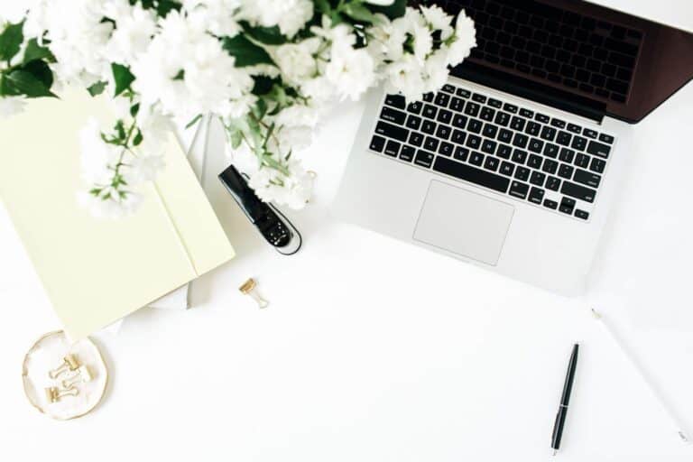 A silver Macbook, with white flowers and stationery on a white desk