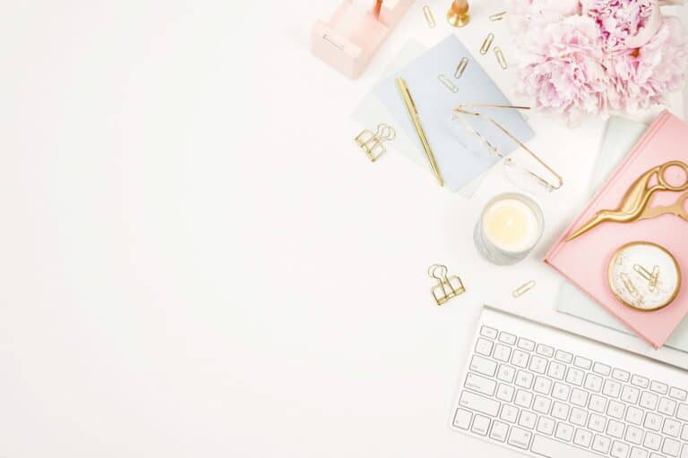 Feminine stationery including a white keyboard, notebooks, golden paperclips and flowers on a white desk