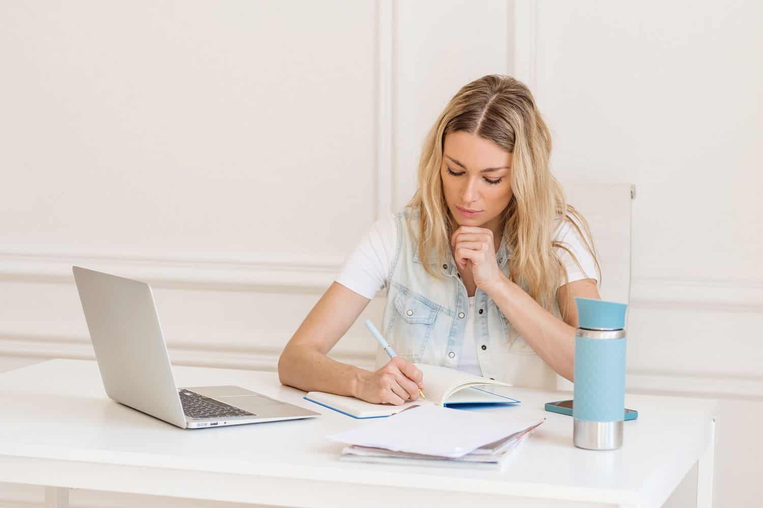 A blond woman sitting at a desk with a silver laptop, writing on a notebook