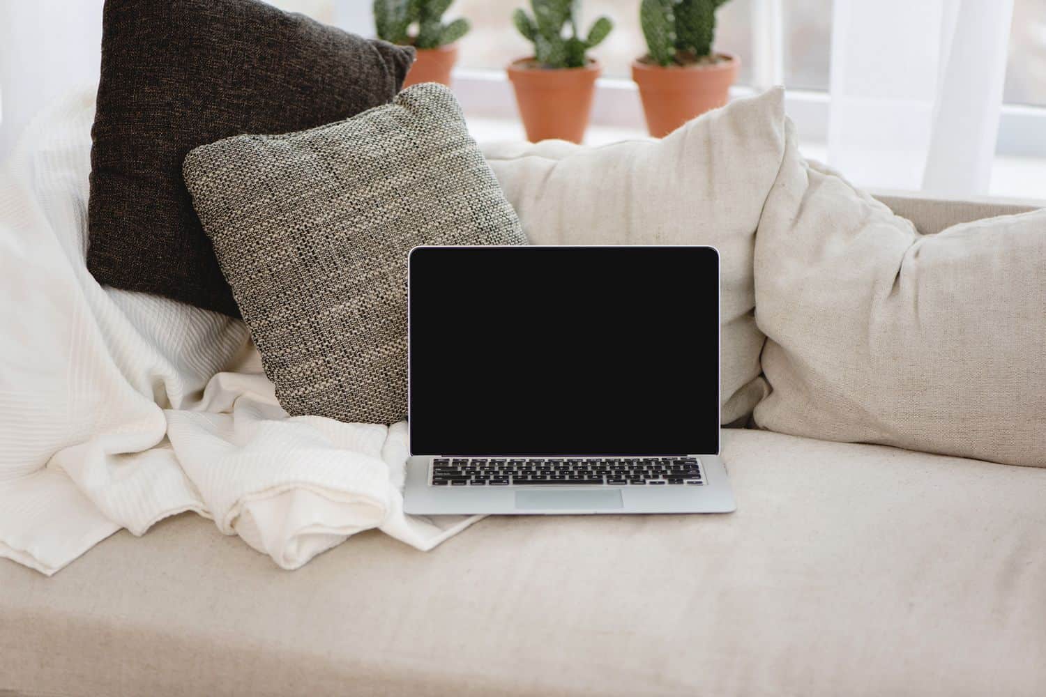 A silver laptop left on a couch with pillows and a blanket