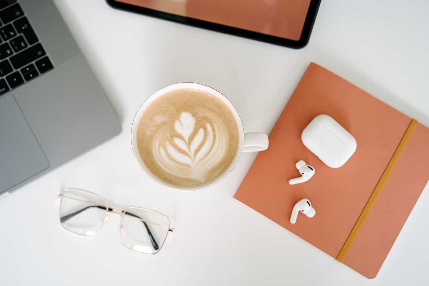 Fall stationery: an orange notebook, lapotop, AirPods, tablet and a latte on a desk
