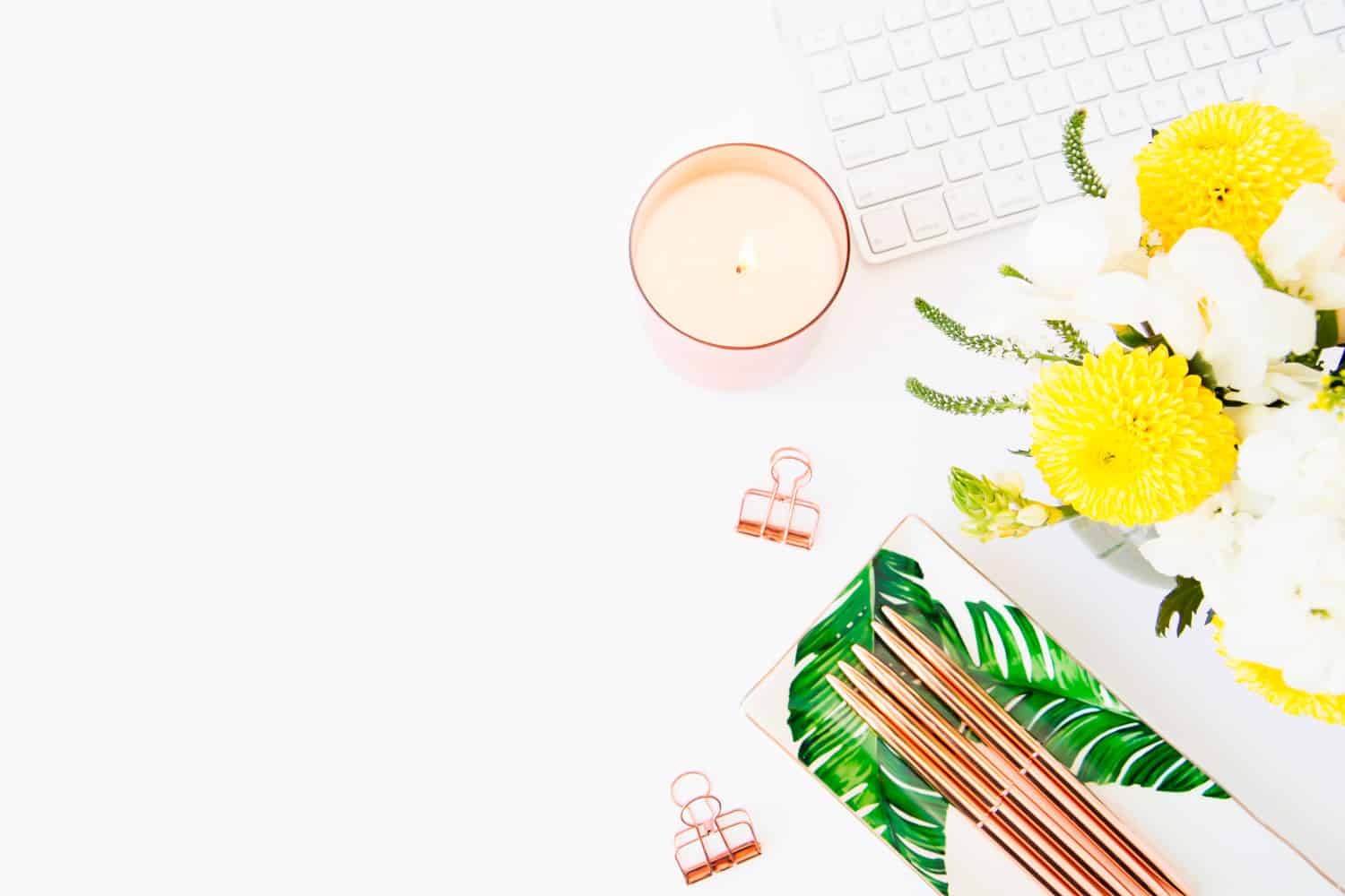 Rosegold pens, yellow flowers, a candle and an Apple keyboard on a desk