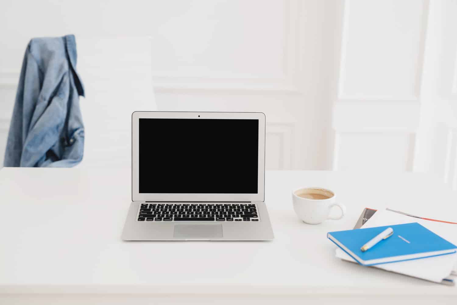 A laptop, mug and notebooks on a table, next to a denim jacket