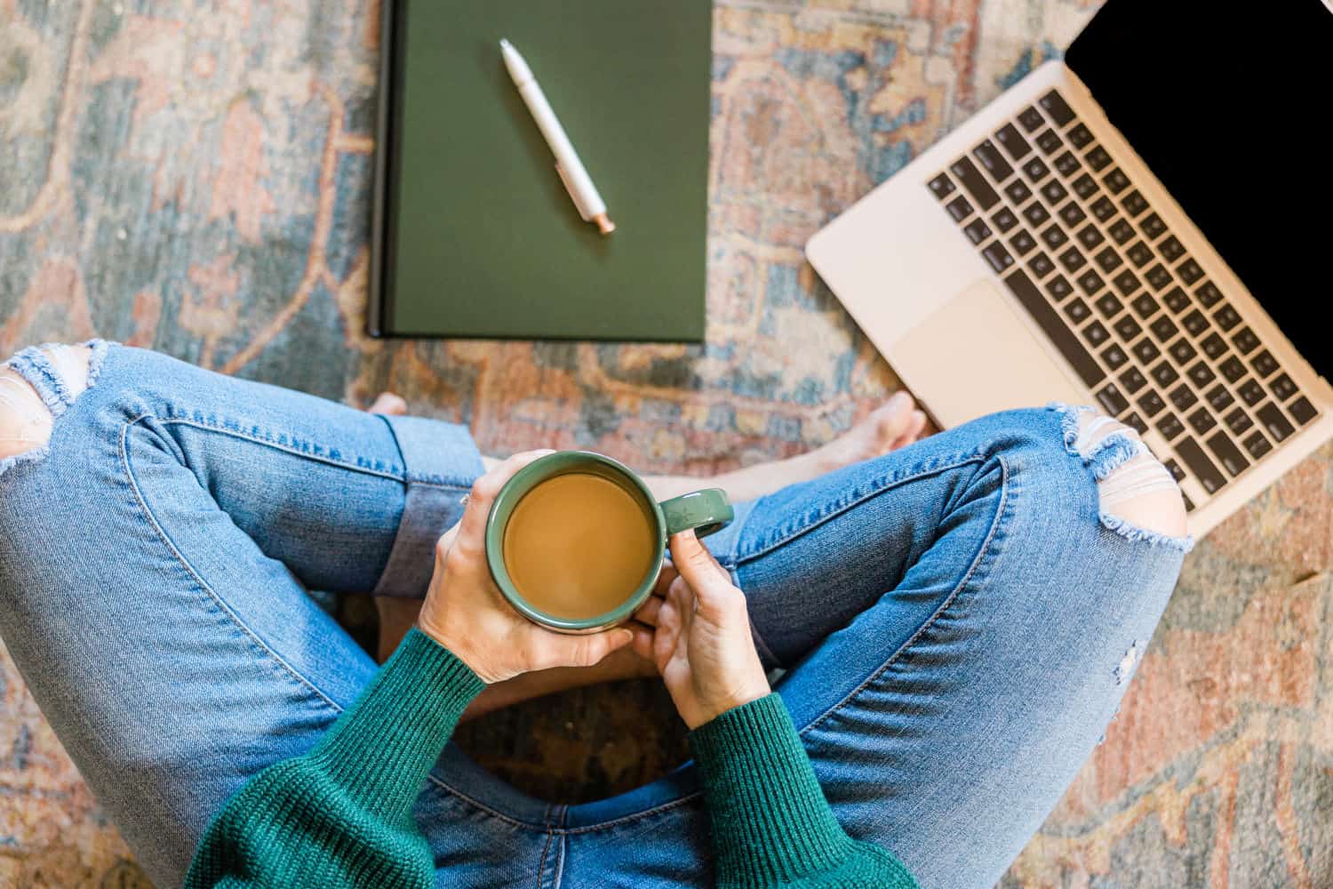 Woman sitting on a carpet next to a laptop and a green notebook, holding a cup of coffee