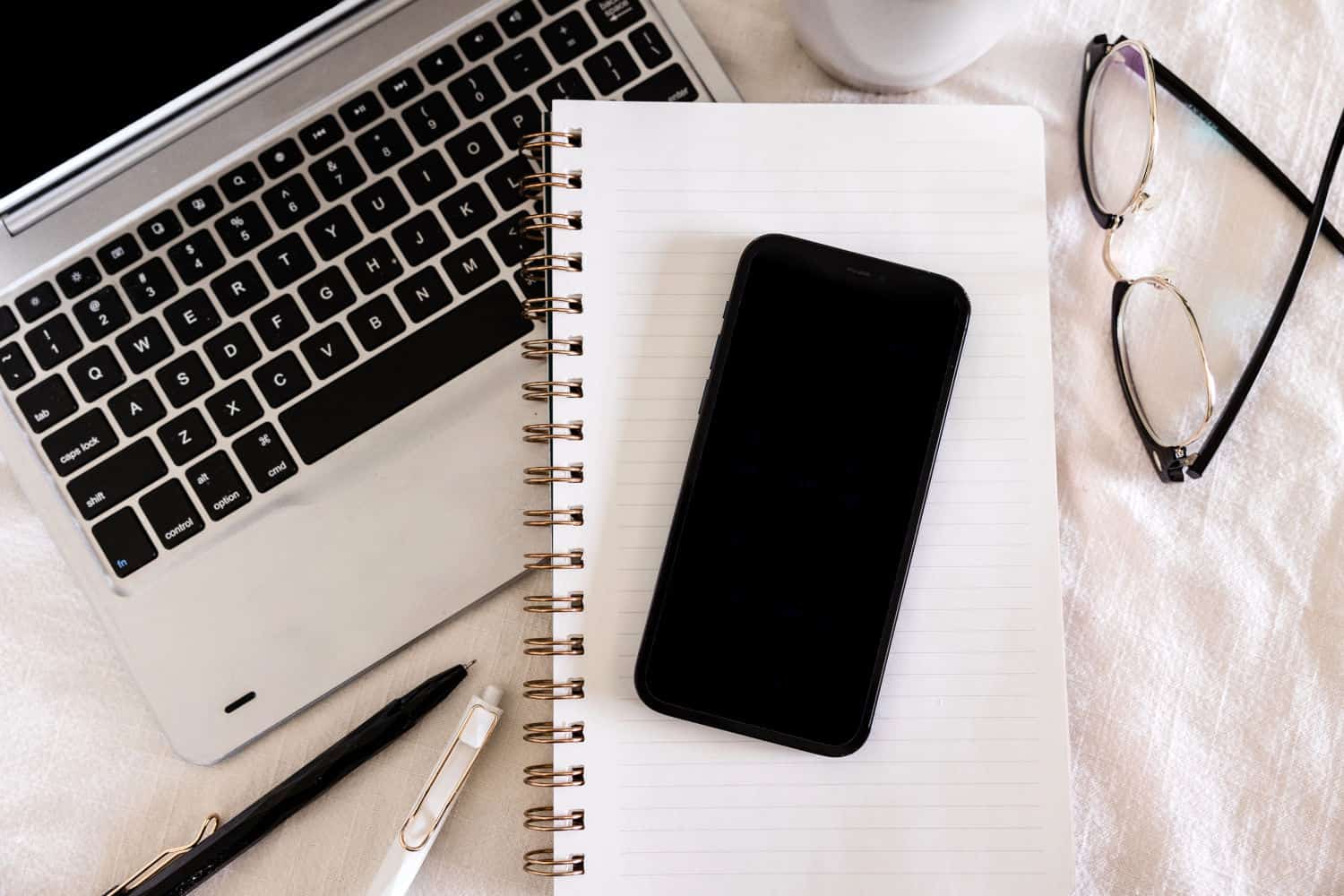 Macbook, open notebook, two pens and a phone on a desk