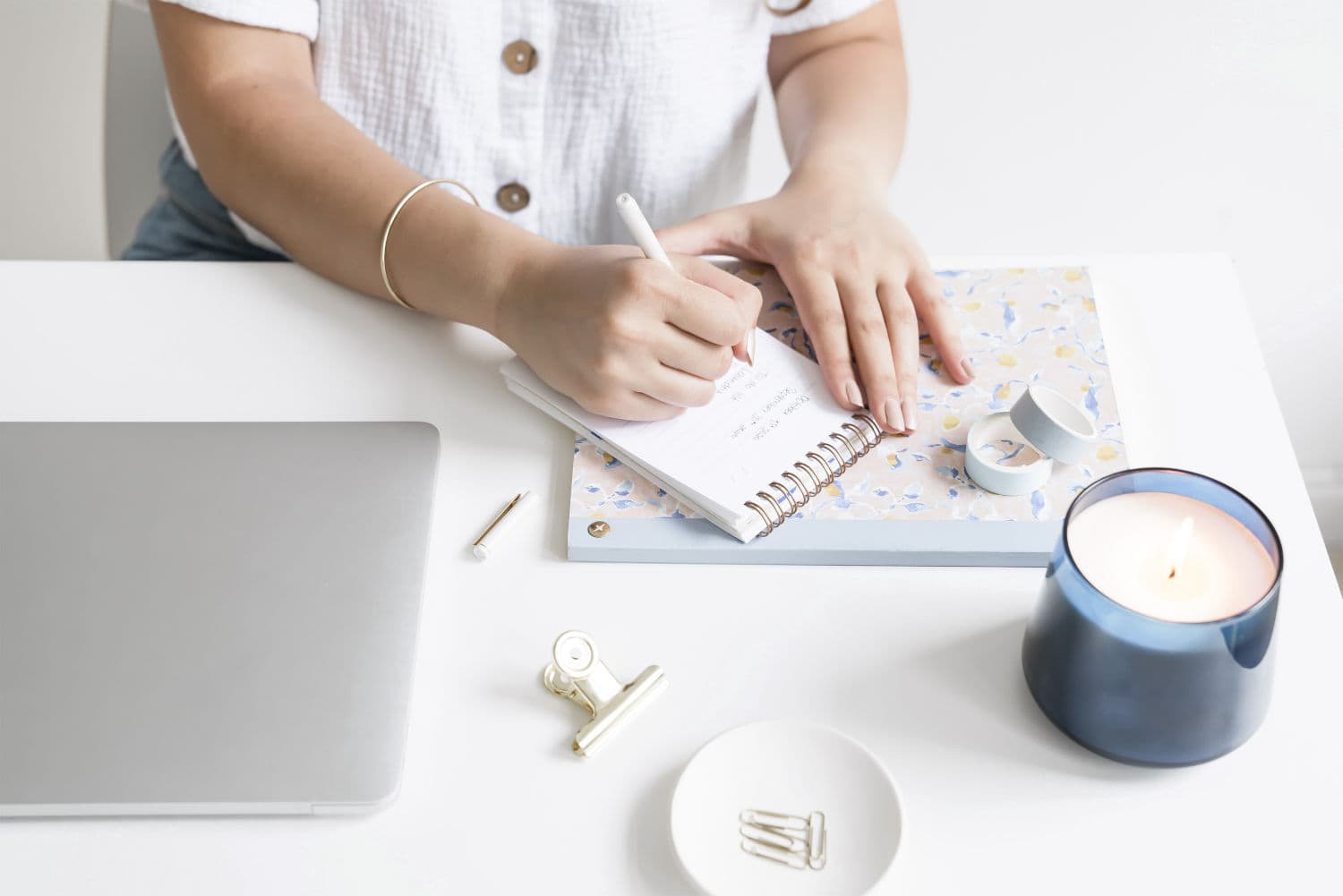 Woman sitting at a desk and writing in a notepad, next to a laptop, paperclips and a blue candle
